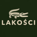 Ciemnozielona koszulka marki Dziedzic Pruski z charakterystycznym autorskim nadrukiem o treści LAKOŚCI i rysunkiem krokodyla tudzież aligatora będacym graficzny nawiązaniem i parodią tenisowych koszulek marki Lacoste.